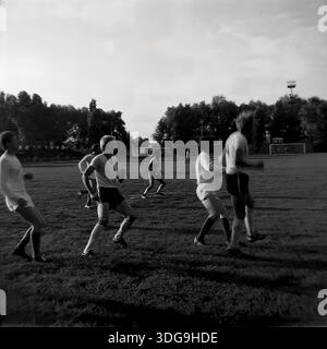 Slawjansk, Region Donezk, Ukraine, UdSSR. Archiv Schwarzweiß-Foto aus den 1980er Jahren, das eine Gruppe von Fußballspielern auf dem Spielfeld des Khimik Stadions zeigt. Die Athleten in Sportuniformen werden in einem dynamischen Moment des Spiels gefangen und laufen über den Rasen. Im Hintergrund bilden dichte Parkbäume und die Stadioninfrastruktur, einschließlich eines Flutlichtturms, die Szene. Dieses Bild veranschaulicht die Beliebtheit der physischen Kultur und den aktiven Lebensstil junger Menschen in der friedlichen Industriestadt vor dem Konflikt. Stockfoto
