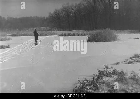 Bezirk Slawjansk, Region Donezk, Ukraine, UdSSR. Archivfoto Schwarzweiß aus den 1960er Jahren mit einer Einzelfigur in einer riesigen Schneelandschaft. Die Person, die in einem dunklen Wintermantel gekleidet ist, hält in der Nähe einer Piste (möglicherweise Skipisten) im Schnee an. Trockene Schilfstellen und hohe Gräser deuten auf ein gefrorenes Sumpfgebiet oder eine Umgebung am Ufer hin. Eine dichte Waldlinie steht im Hintergrund unter einem grau bedeckten Himmel. Dieses stimmungsvolle Bild fängt die Stimmung der Einsamkeit, Stille und die raue natürliche Schönheit der friedlichen Donbass-Region ein. Stockfoto