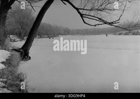 Slawjansk, Region Donezk, Ukraine, UdSSR. Archiv Schwarzweiß-Foto aus den 1960er Jahren mit einer von einem großen Baum umrahmten Winterlandschaft. Ein dunkler, strukturierter Baumstamm lehnt sich von links ab, wobei die Äste nach oben bogenförmig sind und einen natürlichen Rahmen bilden. Durch diese Öffnung ist ein riesiger gefrorener Fluss zu sehen, der mit Schnee bedeckt ist. Winzige Silhouetten von Menschen (wahrscheinlich Fischer) sind auf dem Eis in der Ferne zu sehen. Das gegenüberliegende Ufer ist von Bäumen gesäumt, die in den nebelgrauen Himmel fallen. Dieses stimmungsvolle Bild fängt die Größe und die friedliche Stille der Donbass-Natur ein. Stockfoto