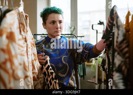 Junge Erwachsene kaukasische Frau, die im Urban Thrift Shop vorgefertigte Kleidung auswählt Stockfoto
