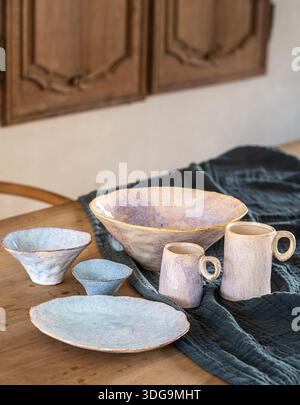 Eine Sammlung handwerklicher Keramikgeschirr, darunter Schüsseln, Teller und Tassen in weichen Lavendel- und Blauglasuren, die auf einem rustikalen Holztisch angeordnet sind. Stockfoto
