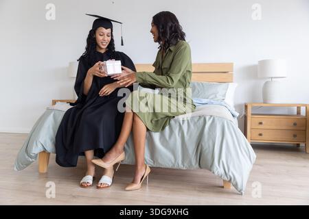 Verschiedene Mutter und Teenager-Tochter tragen Abschlusskittel und Kleid, die auf dem Bett sitzen Stockfoto