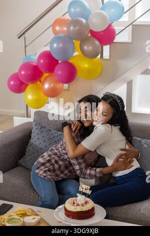 Vielfältige Mutter und Tochter im Wohnzimmer feiern Geburtstag mit Kuchen, Ballons und Stockfoto