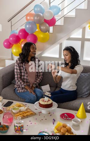 Vielfältige Mutter und Teenager-Tochter sitzen auf dem Sofa und feiern den 18. Geburtstag mit Kuchen Stockfoto