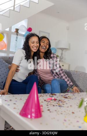 Vielfältige Mutter und Teenager-Tochter umarmen und lachen auf dem Sofa im Wohnzimmer mit Ballons Stockfoto