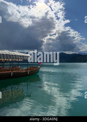 Ein traditioneller Pletna mit gestreiftem Baldachin schwimmt auf türkisfarbenem Wasser unter dramatischen Wolken in Bled, Slowenien Stockfoto