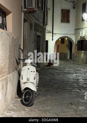 Stimmungsvoller nächtlicher Blick auf eine enge Kopfsteinpflasterstraße in Piran, Slowenien, mit einem klassischen Vespa-Roller Stockfoto