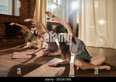 Gruppe junger erwachsener Frauen, die sitzende seitliche Dehnungs-Yoga-Pose auf Matten im sonnendurchfluteten Studio üben, wobei der Schwerpunkt auf Flexibilität und Balance während des Fitnesskurses liegt Stockfoto