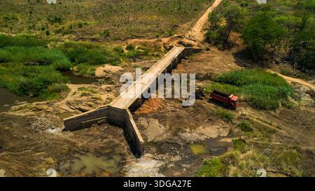 Aus der Vogelperspektive auf eine stabile Betonbrücke über ein flaches, felsiges Flussbett, im Kontrast zur umgebenden grünen Vegetation, Derre, Provinz Zambezia, Moz Stockfoto