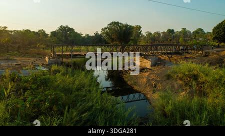 Aus der Vogelperspektive einer robusten Stahlbrücke, die über einen ruhigen Bach führt und den klaren Himmel im üppigen Grün reflektiert, Derre, Provinz Zambezia, Moza Stockfoto