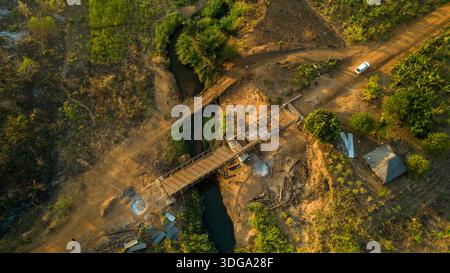 Aus der Vogelperspektive auf eine hölzerne Brücke, die einen schmalen Fluss überspannt, die Landschaft in Gold- und Grüntönen unter der Sonnenumarmung, Derre, Zambezia Provi Stockfoto
