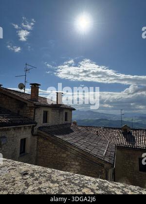 Sonnendurchflutete Dächer eines historischen Steindorfes in San Marino mit blauem Himmel, Wolken und fernen Hügeln, die eine friedliche mittelalterliche Atmosphäre einfangen Stockfoto