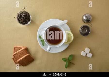 Details einer Teetasse auf einer weißen Keramikplatte auf einem Tisch mit einer braunen Tischdecke mit einer Schüssel mit Teeblättern, Keksen; Minzblätter, eine Scheibe Zitrone, in Stockfoto