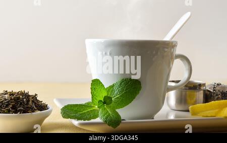 Makrodetail einer Teetasse auf einer Keramikuntertasse mit Minze und Zitrone auf einem Tisch mit einer beigen Tischdecke, Teeblättern und einem Infuser, isoliert auf einem weißen B Stockfoto