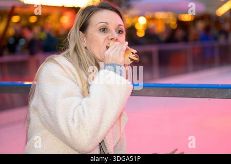 Erwachsene Frau genießt einen Würstchen-Snack auf einem festlichen Weihnachtsmarkt mit bunten Lichtern, winterlicher Atmosphäre und verschwommener Menschenmenge im Hintergrund. Stockfoto