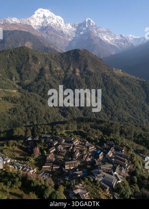 Aus der Vogelperspektive auf ein Dorf eingebettet zwischen den Hügeln, mit schneebedeckten Bergen, die eine atemberaubende Kulisse bieten, Himalaya, Annapurna, Nepal. Stockfoto