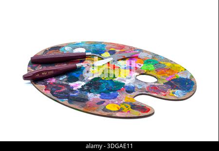 Isoliertes Objekt für Kreativität. Ovale Künstlerpalette aus Holz, bedeckt mit bunten Flecken trockener und frischer Farbe, mit zwei Palettenmessern. Stockfoto