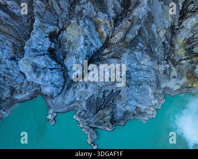 Aus der Vogelperspektive auf das raue Gelände trifft auf das türkisfarbene Wasser, einen starken Kontrast von Texturen und Tönen, Kawah Ijen, Ost-Java, Indonesien. Stockfoto