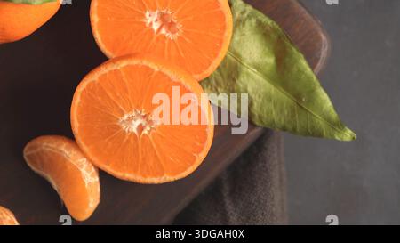 Eine Nahaufnahme von oben nach unten leuchtender Orangenmandarinenhälften, ein einzelnes Segment und ein grünes Blatt, das auf einem dunklen Holzschneidebrett liegt und die festgehalten wird Stockfoto