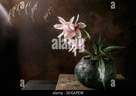 Schöne rosa Magnolienblüten Ikebana in rustikaler grüner Keramikvase vor dunklem strukturiertem Hintergrund. Dramatisches Licht erzeugt stimmungsvolle Atmosphäre Stockfoto