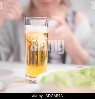 Bier mit Eis und Schaum auf dem Tisch. Ein Glas eiskaltes goldenes Bier aus der Nähe und eine verschwommene Frau im Hintergrund Stockfoto