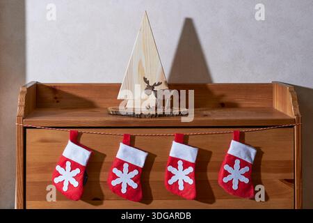 Umweltfreundliche Weihnachtsdekoration. Holzbaum, geschnitzter Hirsch auf Holzscheibe und vier rote Weihnachtsstrümpfe, die an einer Holzkiste hängen. Stockfoto