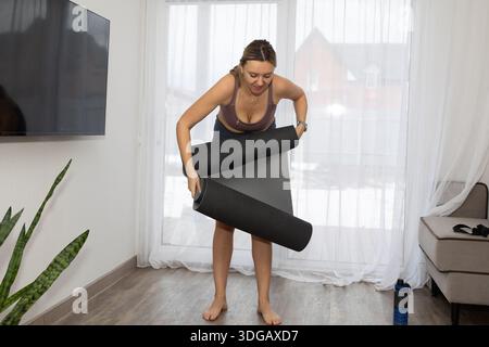 Eine Frau mit Sport-BH und Leggings rollt eine dunkle Yogamatte in ihrem Wohnzimmer auf. Sonnenlicht strömt durch die Vorhänge hinter ihr in der Nähe eines Fernsehers. Stockfoto