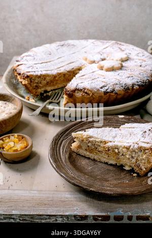Rustikale Ricotta-Tarte Pastiera Napolitano, mit Puderzucker auf Keramikplatte bestäubt. Scheibe auf Holzteller serviert. Kandierte Früchte in einer kleinen Schüssel. Stockfoto