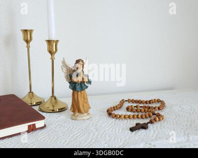Katholische Ecke zu Hause. Engelsfigur, Rosenkranzperlen aus Holz, Kreuz und Gebetsbuch. Glaube, Symbole und Traditionen. Stockfoto