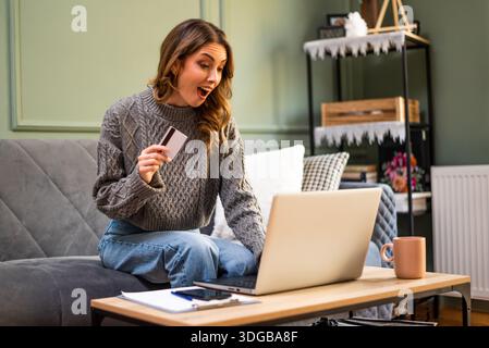 Glückliche Erwachsene Frau hält eine Zahlungskarte in der Hand und benutzt einen Laptop, um Rechnungen oder Waren online zu bezahlen. Stockfoto