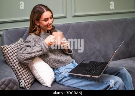 Eine lächelnde Frau entspannt sich auf einem Sofa mit einem warmen Getränk in der Hand und arbeitet an ihrem Laptop. Stockfoto
