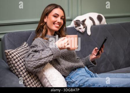 Glückliche Erwachsene Frau entspannt sich zu Hause. Sie sitzt auf dem Sofa, trinkt Kaffee und benutzt das Smartphone, während die Katze neben ihr liegt. Stockfoto
