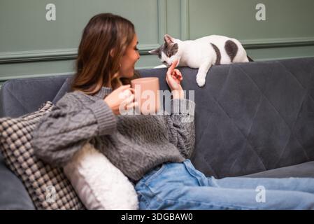 Glückliche Erwachsene Frau entspannt sich zu Hause. Sie sitzt auf dem Sofa und trinkt Kaffee, während die Katze neben ihr liegt. Stockfoto