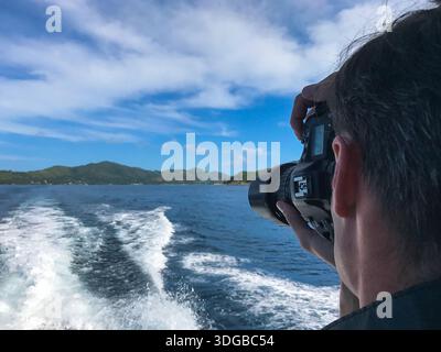 Rückansicht eines Mannes, der eine DSLR-Kamera hält, während er Meereslandschaften von einem fahrenden Boot aus fotografiert. Sanfte Wellen und fließendes Wasser im Vordergrund. Stockfoto