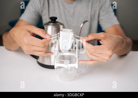 Nahaufnahme des zugeschnittenen Bildes eines nicht erkennbaren Mannes, der einen Tropfkaffeesack auf eine Glasschale legt und den Prozess des Zubereitens frisch gemahlenen Kaffees mit dem Wasserkocher zeigt Stockfoto