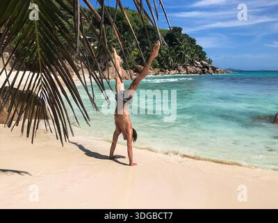 Sportlicher Mann, der einen Handstand mit auseinander liegenden Beinen an einem tropischen Strand mit weißem Sand und türkisfarbenem Wasser macht. Palmblätter umrahmen die Szene, mit grüner Küste. Stockfoto