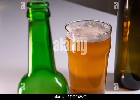 Ein Atelier aus der Nähe eines mit Bernsteinbier gefüllten Glases mit einem schäumenden weißen Kopf neben einer verschwommenen grünen Flasche auf weißem Hintergrund. Stockfoto