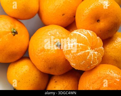 Eine Nahaufnahme von oben nach unten auf einen Haufen frischer Mandarinen. Der Großteil der Früchte ist ganz mit hellorange, strukturierter Haut, während eine Mandarine geschält wird Stockfoto