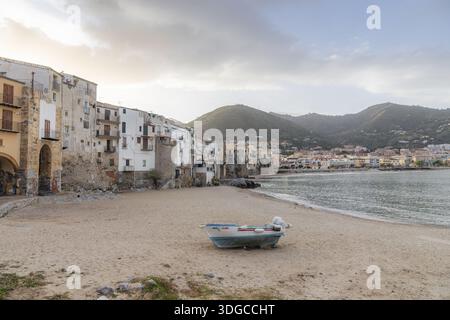 Blick auf die verwitterten Gebäude an der Küste, wo ein kleines Boot am Sandstrand unter weichem Himmel liegt, Cefalu, Sizilien, Italien. Stockfoto