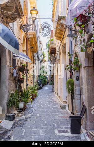 Blick auf eine malerische gepflasterte Gasse, gesäumt von alten Gebäuden, Topfpflanzen und dekorativen Regenschirmen, die Schatten in Ortigia, Sizilien, werfen. Stockfoto