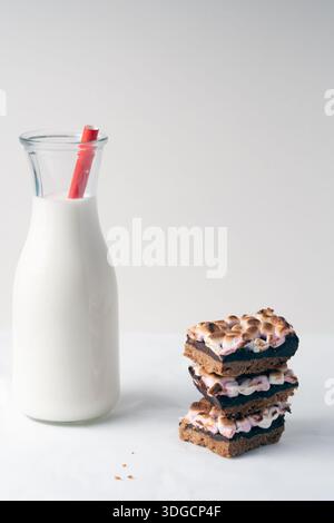 Ein Glas Milch sitzt neben einem Stapel Brownies. Die Brownies sind mit Marshmallows auf hellem Hintergrund bedeckt Stockfoto