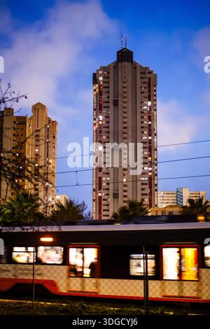 Eine lebendige Nachtaufnahme eines Zuges, der durch Benidorm fährt, mit der Bewegung der Autos im Kontrast zu den statischen, hell beleuchteten Wohntürmen. Stockfoto