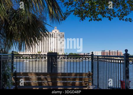 Eine Bank sitzt hinter einem Eisengeländer mit Blick auf den Savannah River und gegenüber der Uferlinie des Gebäudes im historischen Baumwoll-Lagerviertel in Sav Stockfoto