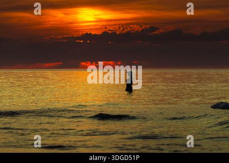 Silhouette einer Person, die während des dramatischen Sonnenuntergangs in Łeba, Polen, in ruhigem Meerwasser steht. Stockfoto