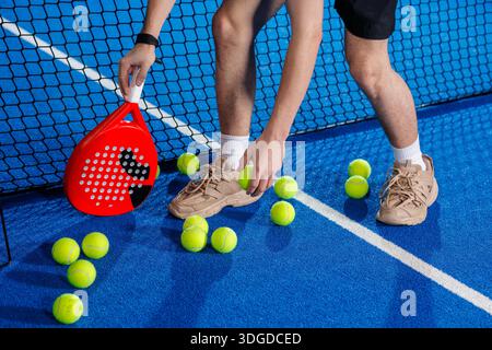 Der Spieler sammelt während der Trainings- und Übungseinheit Padelbälle neben dem Netz auf einem Hallenplatz Stockfoto