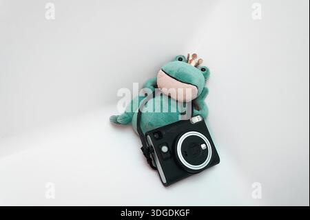 Niedlicher Plüschfrosch mit Krone, chillt mit einer sofortigen Kamera auf weißem Hintergrund, minimales kreatives Konzept für Fotografie und lustiges Design. Stockfoto