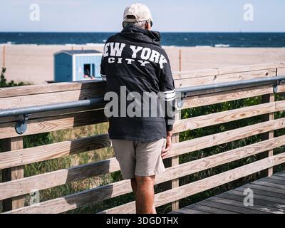 Ein Mann mit einer New York City Jacke steht an der Promenade und blickt an einem klaren Tag auf den Atlantik. Stockfoto