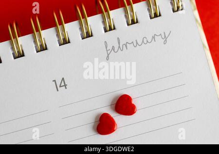 Nahaufnahme eines Kalenders am 14. Februar und zwei kleine rote Herzen auf rotem Hintergrund. Valentinstag-Festkonzept mit Kopierraum. Stockfoto