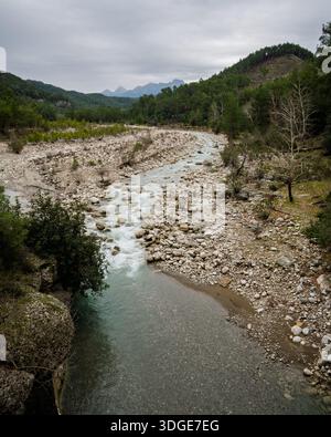 Malerische Landschaft mit einem trockenen, breiten Flussbett mit weißen Steinen, umgeben von grünen Kiefernwäldern und Bergen an einem bewölkten Tag. Stockfoto