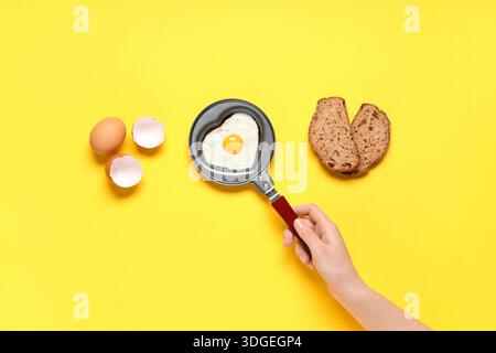 Eierschale, Stück Brot und weibliche Hand mit herzförmigem Spiegelei in der Pfanne auf gelbem Hintergrund Stockfoto